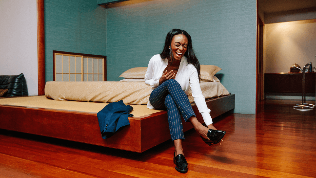 A woman wearing work attire, laughing while fixing her shoe in a teal bedroom