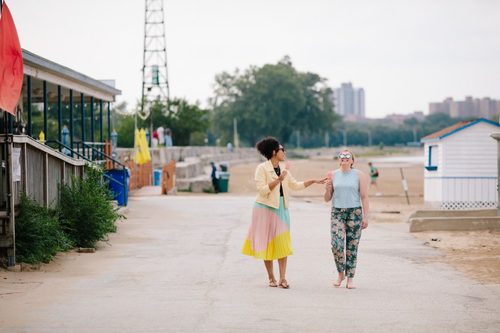 Two women walking on a sidewalk and eating popsicles.
