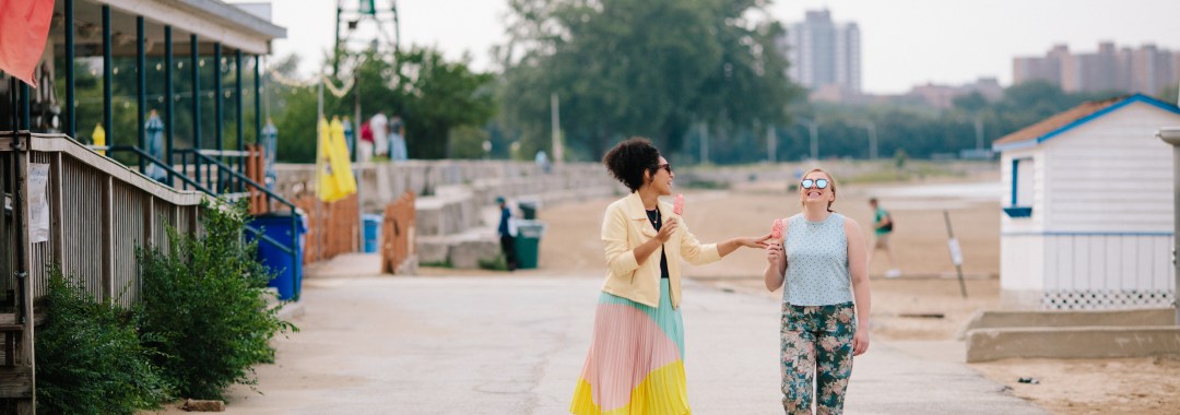 Two women walking on a sidewalk and eating popsicles.